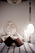 Weisses Gipsrelief im antiken Stil und eiförmiges Fensterchen hinter Deko-Kissen und Tagesdecke in Naturtönen