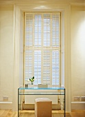 Simple, glass desk with shelf and leather stool in front of closed window shutters in high-ceilinged room in old building