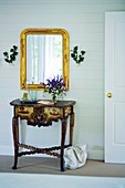 Antique, carved console table below gilt-framed mirror in rustic foyer