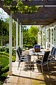 Table and chairs beneath pergola with climbers adjoining country house