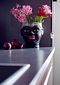 Blumengefäss in Gestalt eines schwarzen Frauenkopfes mit Sommerblumen und rote Zwiebeln auf Küchenzeile