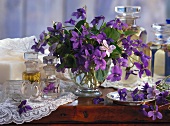 Purple spring bouquet in glass vase next to various glass containers on wooden shelf