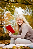 Young woman reading book on blanket in garden with autumnal foliage