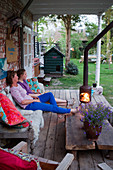 Mutter und Tochter sitzend auf rustikaler Holzbank vor Feuerstelle auf Terrasse