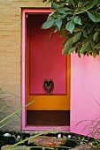 Blick von Aussen durch hellviolette Tür auf pink und orange gestrichene Wandnische mit Tierkopf in stilisiertem Herz
