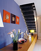 Blumenstrauss auf Sideboard aus Holz vor kobaltblau getönter Wand und Blick auf schwarze Untersicht des gefalteten Treppenlaufes. Modernes Ambiente.