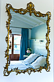 Spiegel mit Goldrahmen und floraler Verzierung an Wand und gespiegeltes Bett vor Fenstertüren mit blauem Vorhang