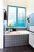 A built-in bath under a large window with blinds with a wooden shelf and a vase of flowers
