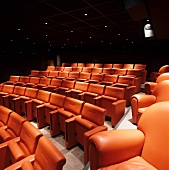 Kinosaal - Stuhlreihen mit orangefarbenen Rücken- und Sitzpolster unter dunkler Kassettendecke mit Strahlern