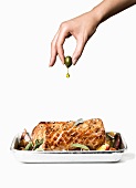 A hand squeezing olive oil from an olive onto roast pork with apple and sage
