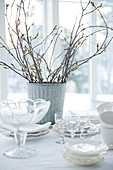 Dishes and glasses in front of metal vase of twigs on table with tablecloth