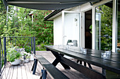 Rustic, black-painted, long table and bench on wooden terrace with open terrace doors; summer greenery in background