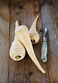 Three parsnips on a wooden surface with a peeler