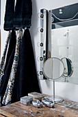 Modern bathroom mirror with row of lights on one side, small silver boxes on old table and black towels hanging on wall