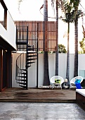 Stylish courtyard of contemporary house with white wicker chairs on wooden platform in front of palm trees and spiral staircase