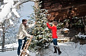 Junges Paar dekoriert schneebedeckten Christbaum vor einer Almhütte