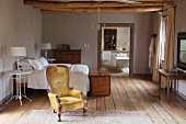 Antique armchair with gold upholstery in front of double bed in rustic setting with view into bathroom through open doorway