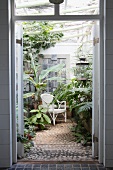 View through open door into conservatory with tropical plants and white wicker chair on cobbled floor