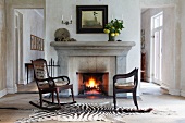 Vintage rocking chair and antique armchair on zebra-skin rug in front of open fire in simple house