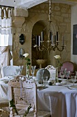 Messing-Kerzenleuchter über festlich gedeckter Tafel mit antiken, weissen Stühlen im Vintage-Look; Natursteinwand im Hintergrund