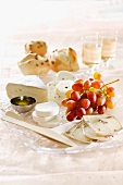 A cheese platter with grapes, rolls and wine in the background