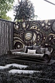 Ausladendes Korbsofa vor Rückwand mit psychedelischem Schwarzweiss-Muster, darüber ein aufgehängtes Kunstobjekt aus kleinen Ästen