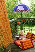 Exotic seating area on balcony with blue parasol with beaded fringe and wedge cushions on colourful blanket