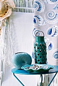 Mint green ceramic lantern on small side table; straw hat and curtain with shell design in background