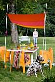 Selbstgemachtes Sonnensegel an abgespannten Bambusstäben über gedecktem Tisch mit orangefarbenen Sesseln und Dalmatiner; Frau unscharf im Garten-Hintergrund