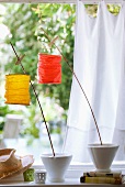 Kleine Papierlampions, mit Wäscheklammern an Holzstäben befestigt, in Gartenatmosphäre mit weißem Vorhang im Hintergrund