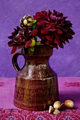Dunkelrote Dahlienblüten in klassischem Keramikkrug vor violettem Hintergrund; Eicheln auf bestickter Leinendecke