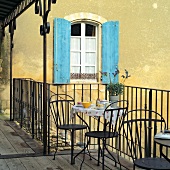 Metal bistro table set for two on wooden balcony with simple metal railing; blue shutters in background
