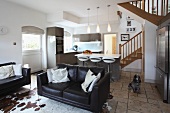 Leather sofas and cow-skin rug in open-plan interior with modern fitted kitchen and country house style staircase in background