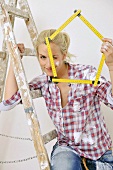 Renovierungsarbeiten - Frau sitzt auf der Leiter und hält einen Meterstab in der Hand