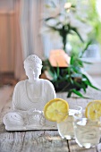 Refreshing drinks next to seated Buddha figurine in white stone on wooden floorboards