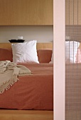 Double bed with pale bedspread against smooth wooden fronts of fitted cupboards; detail of sliding door with translucent bamboo blind