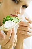 Young woman holding up cracker topped with cucumbers and cheese spread, cropped