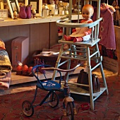 Alte Puppe in raffiniertem Kinderkombistuhl aus alten Zeiten und blau lackiertes Vintage Dreirad in Secondhand Boutique
