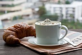 A cappuccino with a croissant on newspaper in front of a window