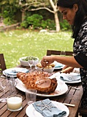 A woman carving larded roast ham on a table in a garden