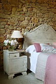 Bedside cabinet painted light grey next to bed with headboard of same colour against stone wall