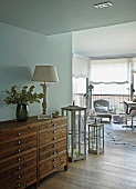 Half-height, wooden vintage chest of drawers against wall in open-plan living room with view of armchair in front of windows with half-opened blinds