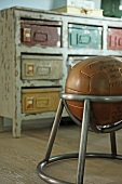 Leather ball in metal chair frame in front of vintage chest of drawers with metal boxes in place of drawers