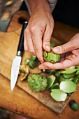 Woman's Hands Peeling Baby Artichokes Over a Wooden Cutting Board