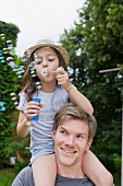 Girl sitting on father's shoulders blowing soap bubbles