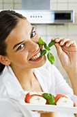Woman biting into sprig of basil, holding plate of Caprese salad