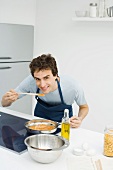 Man cooking in kitchen, bending over to taste sauce, smiling at camera