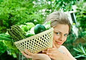 Woman holding basket full of fresh vegetables, smiling at camera