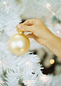 Goldene Weihnachtskugel in der Hand haltend vor künstlichem Weihnachtsbaum
