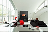 View across stainless steel kitchen counter to open-plan interior with black sofa and red designer armchair next to huge window